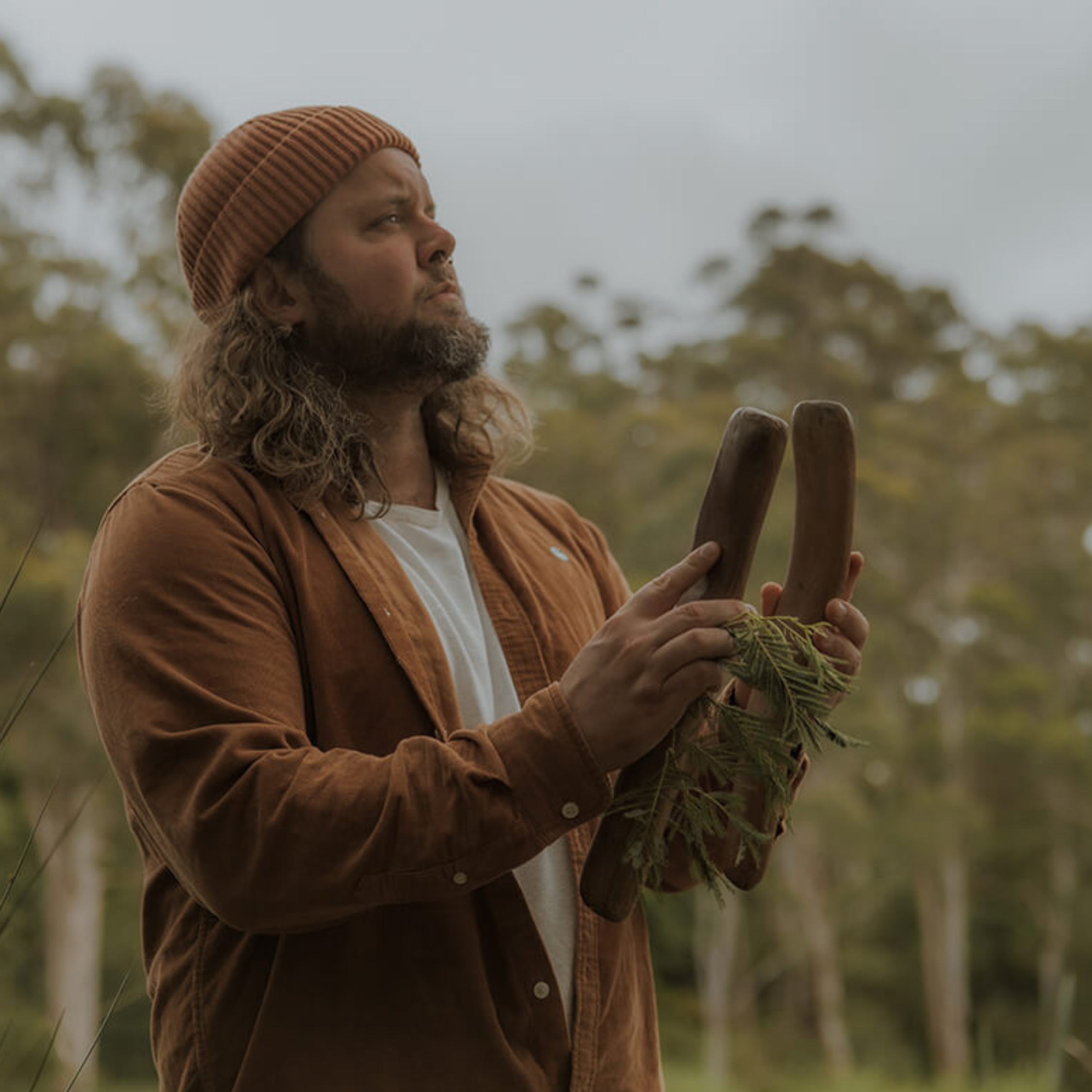 An Indigenous Australian man stands in bushland holding two carved wooden sticks wrapped with native leaves.