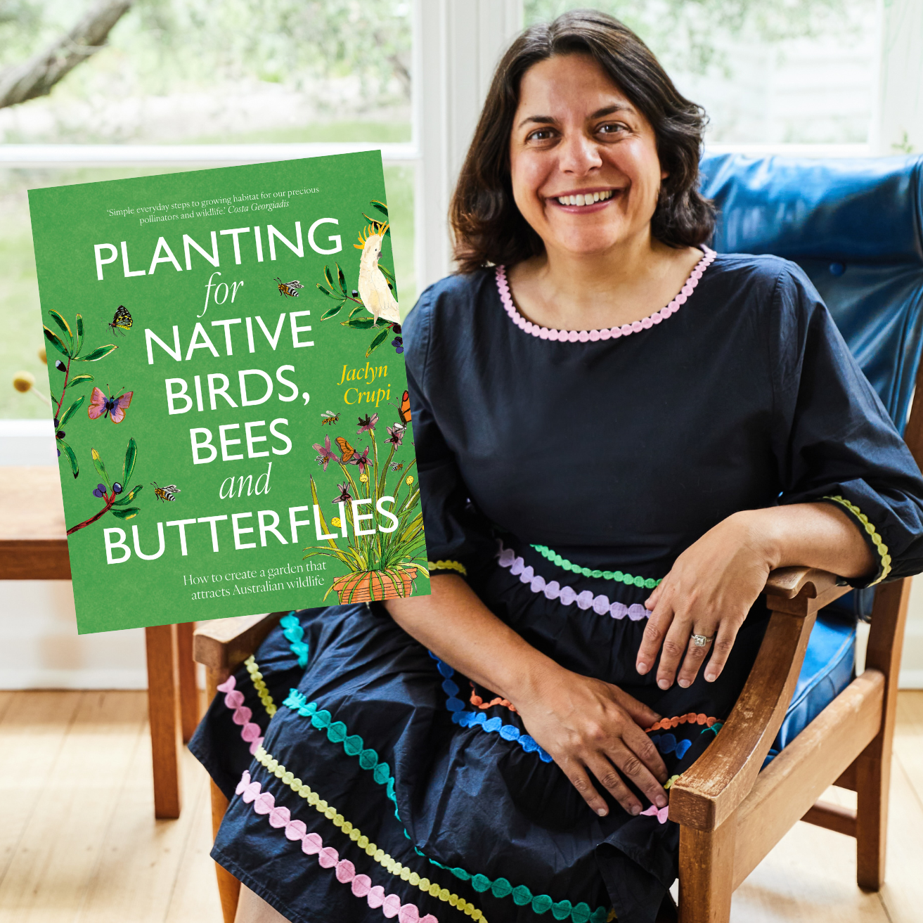 A lady in a blue dress with shoulder length dark hair is looking at camera and smiling. She is seated in front a window and looks relaxed and happy. Next to her is a book titled Planting for Native Birds, Bees and Butterflies by Jaclyn Crupi.