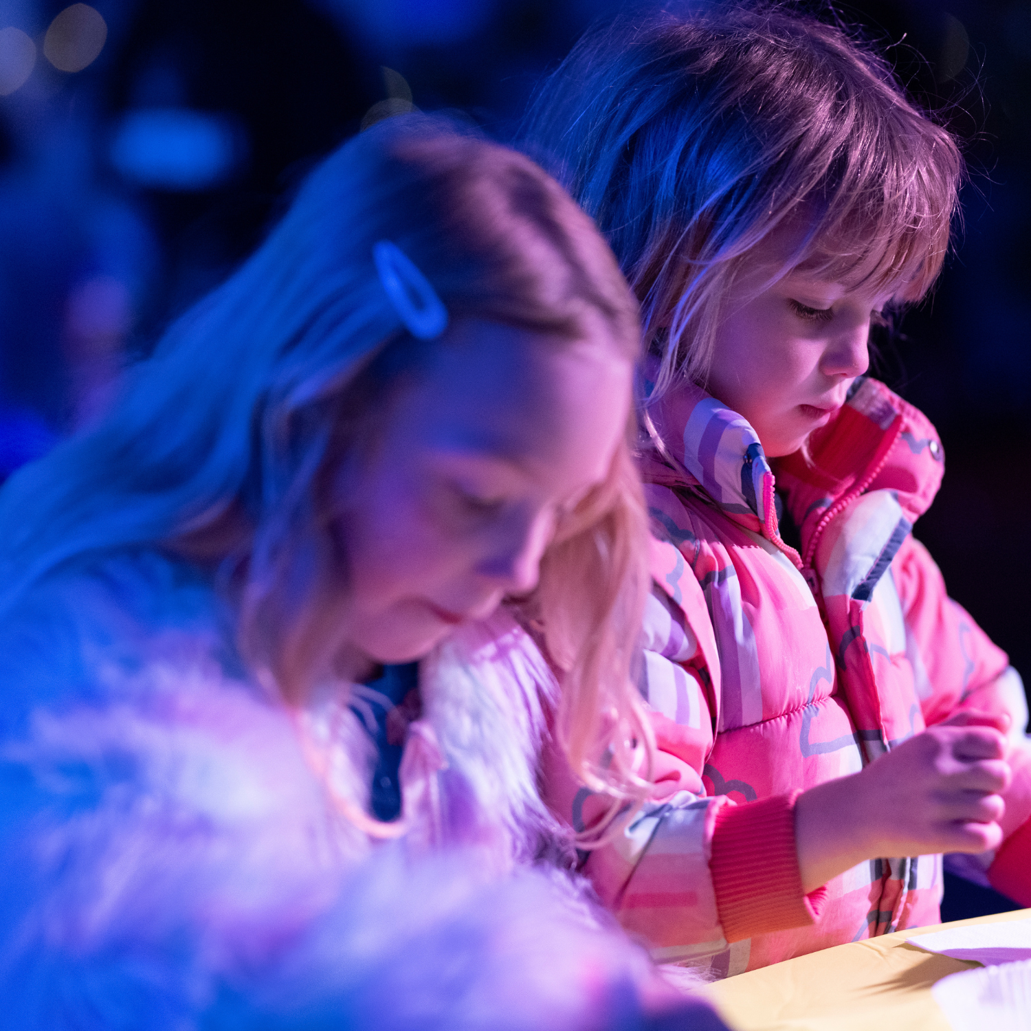 Two children in winter coats in a darkened setting creating something craft like at a table.