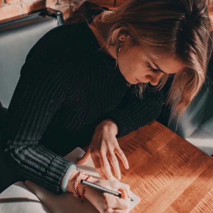 A woman in a black jumper is writing a letter at a table. 