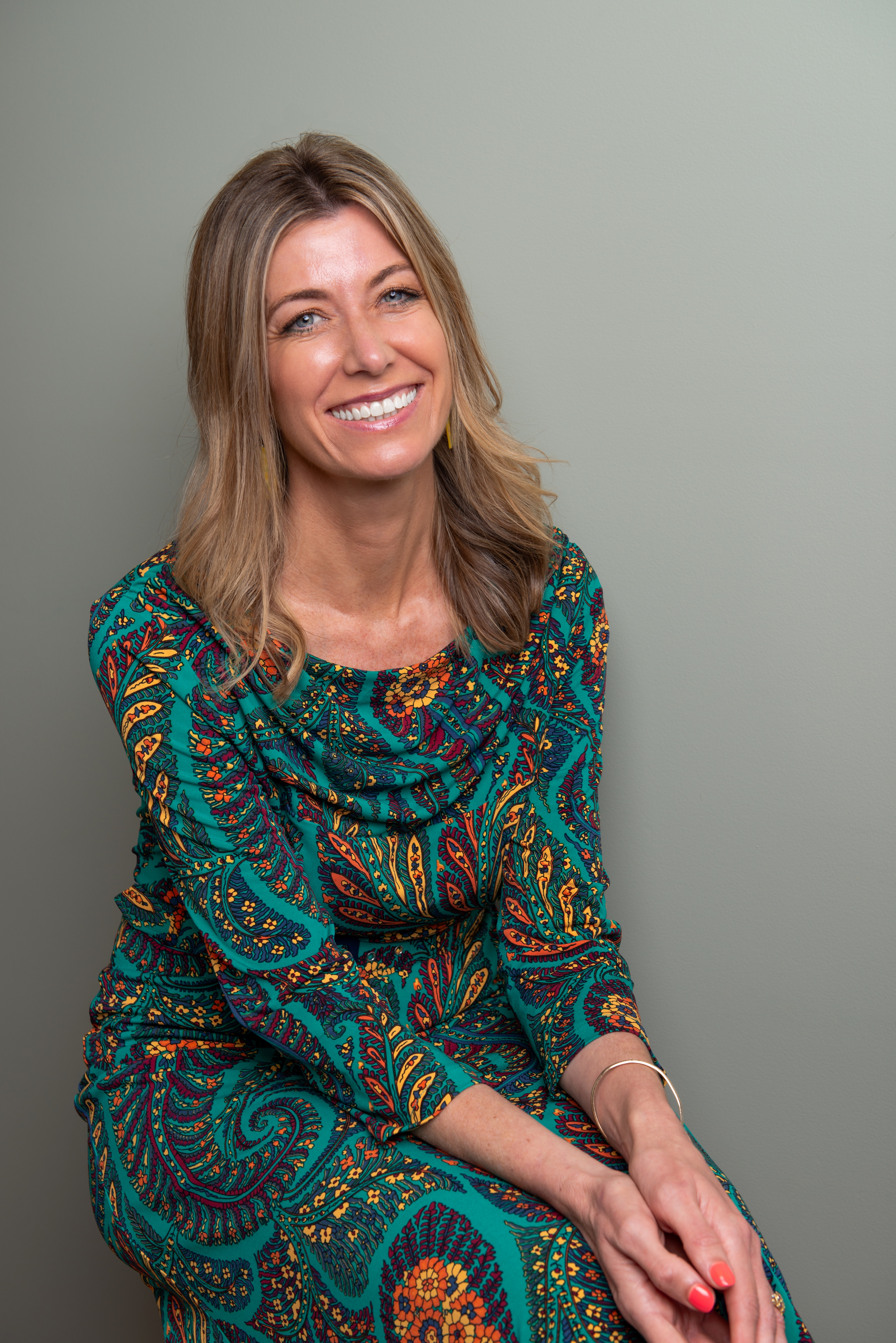 A lady with blonde hair and a big smile is looking at the camera. She is seated with her hands folded on her lap. She is wearing a bright colourful dress.