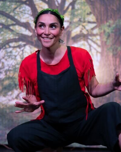 A lady in overalls crouching, looking to her left and smiling. A tree backdrop is behind her.