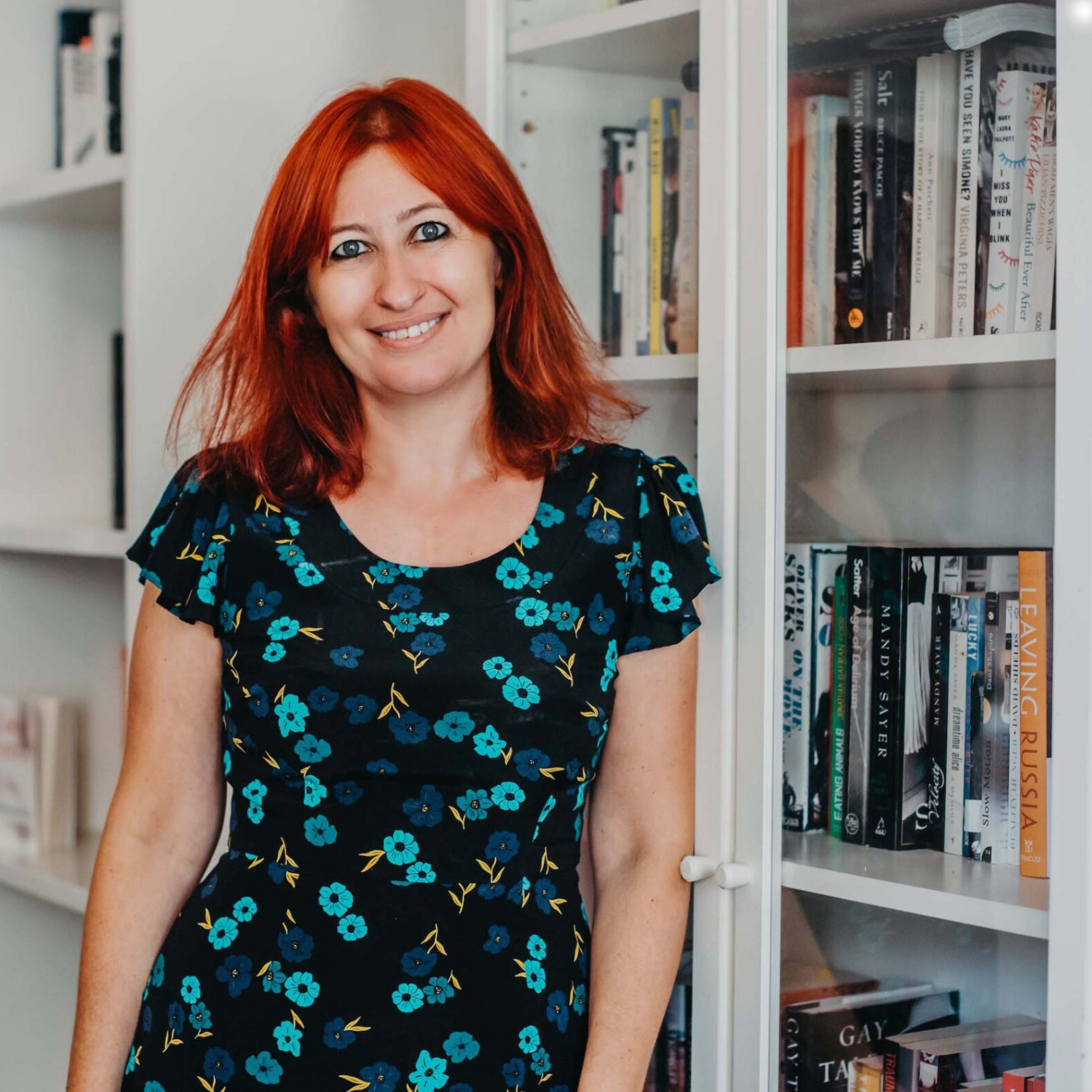 A woman with red hair and a floral dress looks at the camera. She is standing in front of a bookshelf.