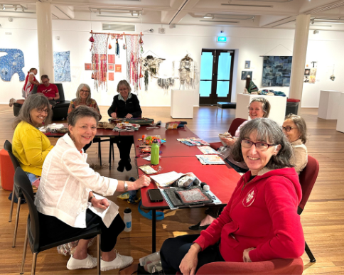 A group of women looking at the camera while sitting at a table. The table is full of craft supplies. They appear to be sitting inside a gallery exhibition space.