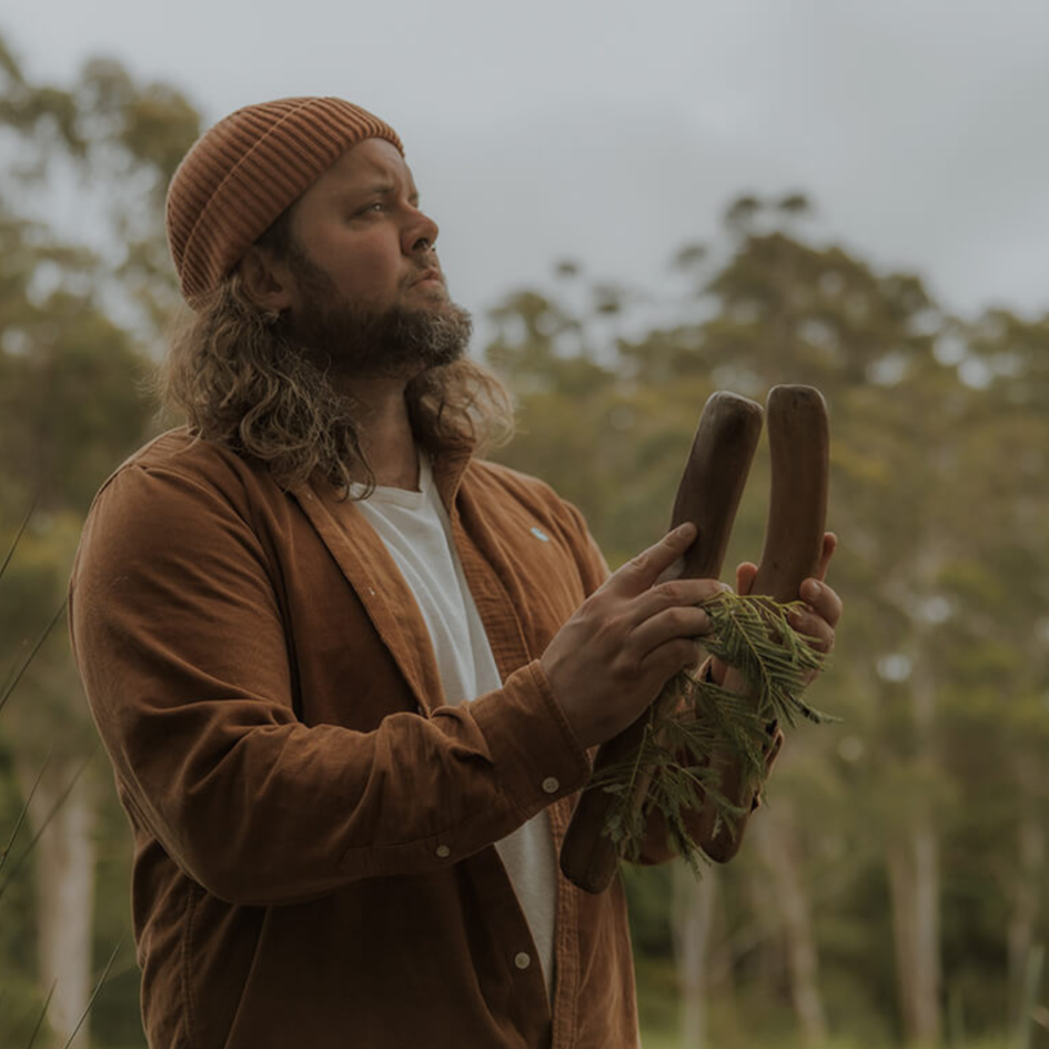 An Indigenous Australian man stands in bushland holding two carved wooden sticks wrapped with native leaves.