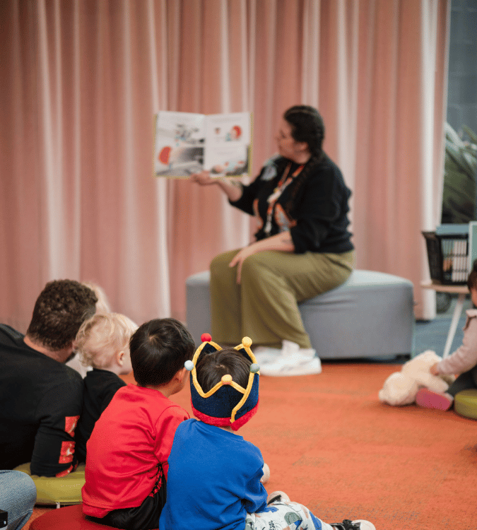 A library staff member reads a picture book to a group of young children and adults seated together during a Storytime session.