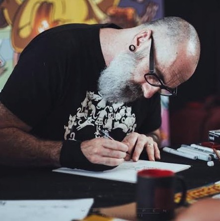 Dean Rankine, comic book artist and illustrator, sits at a table concentrating as he sketches in a notebook.