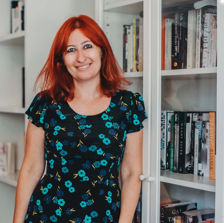 a lady with red hair and a floral dress is smiling at the camera. She is standing in front of a bookshelf.