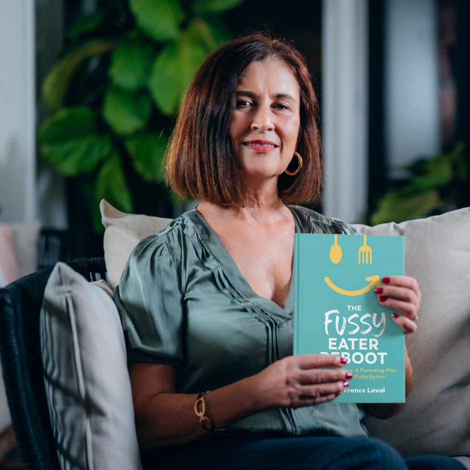 Woman seated on a sofa, smiling and holding a copy of The Fussy Eater Reboot by Marie France-Laval.