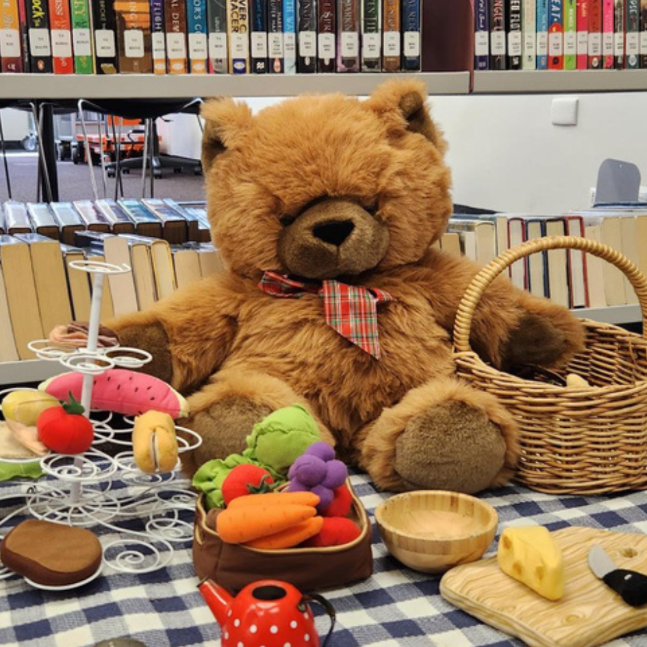 A large teddy bear sits on a rug in the library surrounded by toy tea party items. 