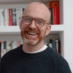 A man with glasses and a beard smiling at the camera. There is a bookshelf behind him.