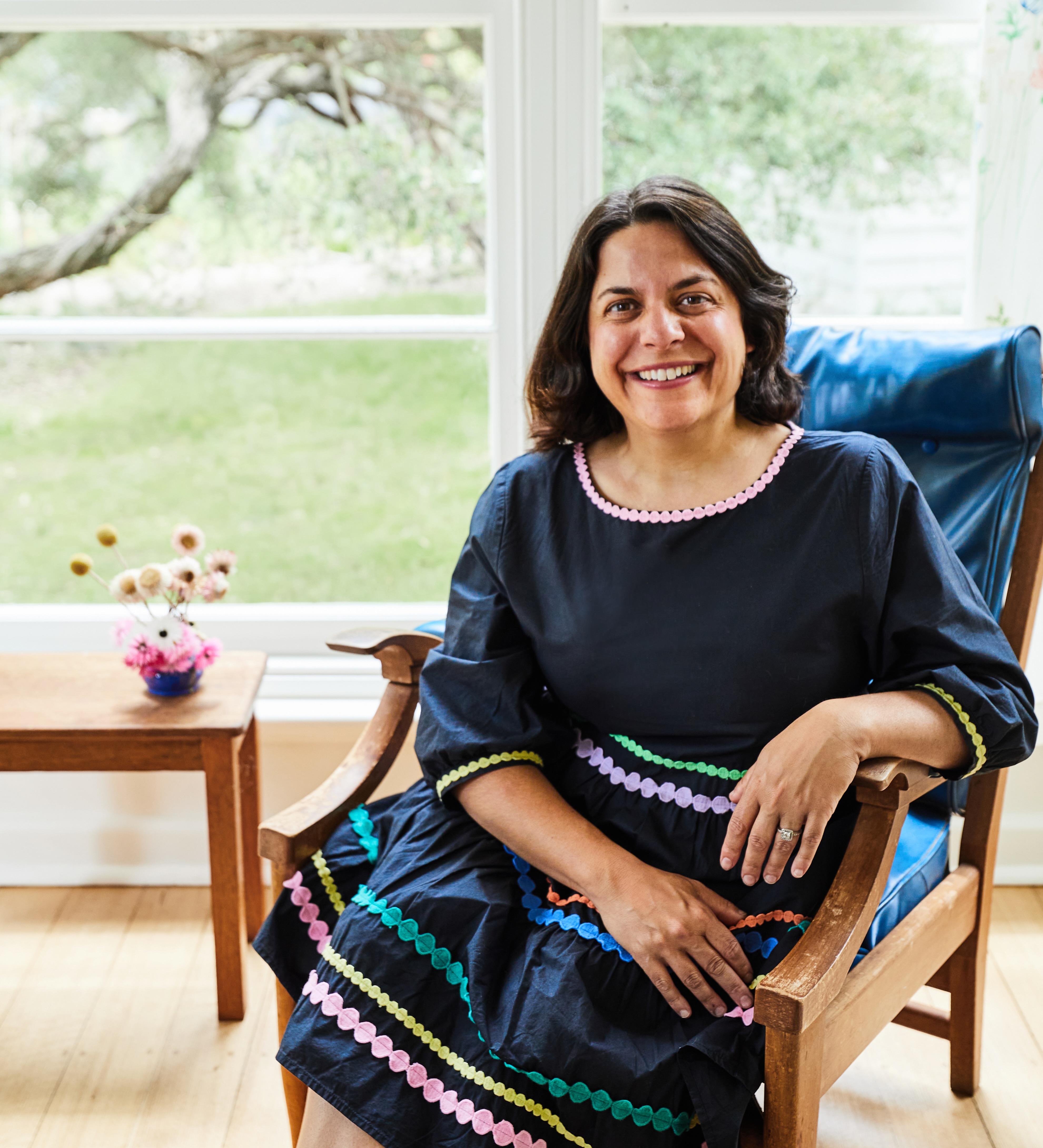A lady with short black hair sitting in a chair in front of a window. She is wearing a blue dress and looks relaxed. She is smiling.